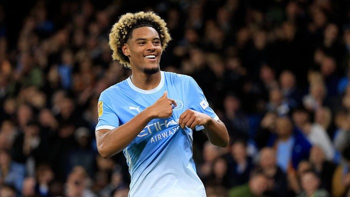 Footballer in a sky-blue Manchester City jersey celebrates on the field with a thumbs-up gesture as the crowd looks on in the stands.