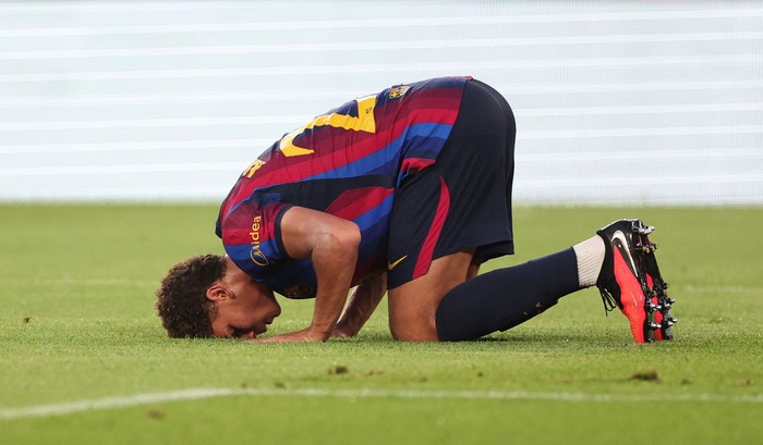 Soccer player in a Barcelona jersey kneels and bends forward with hands on the grass on a stadium field, wearing red-and-black kit and bright cleats.