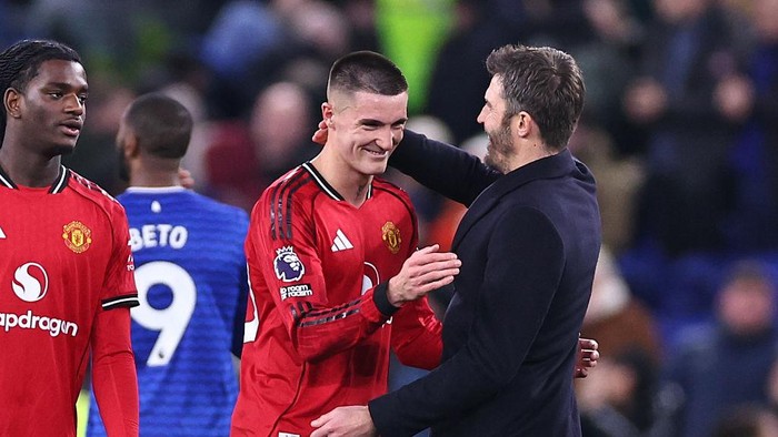 Two Manchester United players in red shirts shake hands with a man in a dark coat on the pitch, with a blue-shirted opponent in the background.