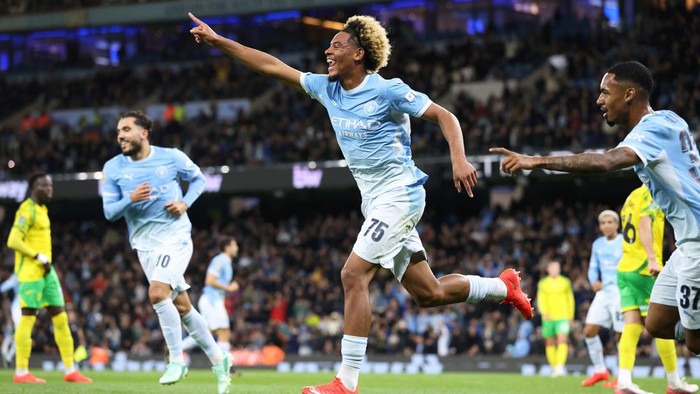 Manchester City players celebrate a goal on the soccer field as fans cheer in the stands, with players in light blue uniforms running and pointing.