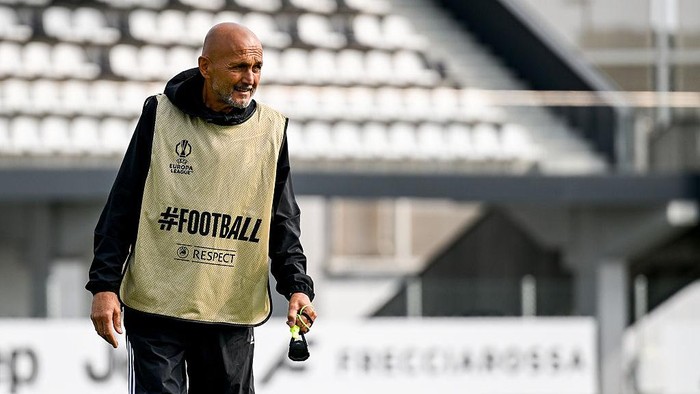 Bald man wearing a beige training bib with '#FOOTBALL' and 'RESPECT' on a soccer field, holding a whistle.