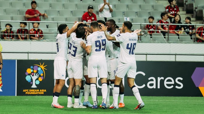 Soccer players in white uniforms form a huddle to celebrate on the green field, with fans watching from the stands behind them.