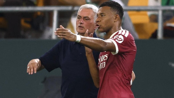 Coach in a dark shirt speaks and gestures while a player in a maroon jersey points ahead during a soccer match.