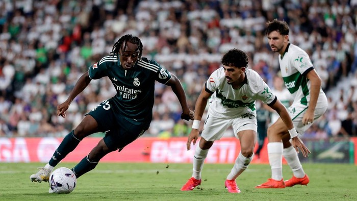 Soccer player in dark jersey dribbling the ball while two opponents in white-green kits chase nearby on a green field during a crowded match stadium.