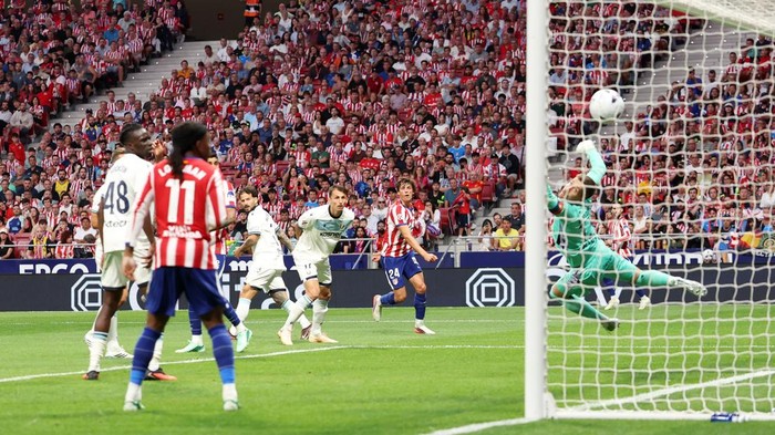 Soccer match scene: players in white and red stripes near the goal as the ball heads toward the net, with a goalkeeper in green diving to make a save and a crowded stadium backdrop.
