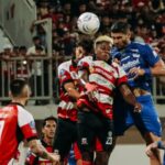 Soccer players from red-white and blue teams leap to head the ball amid a crowded box in a match.