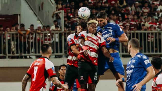Soccer players from red-white and blue teams leap to head the ball amid a crowded box in a match.