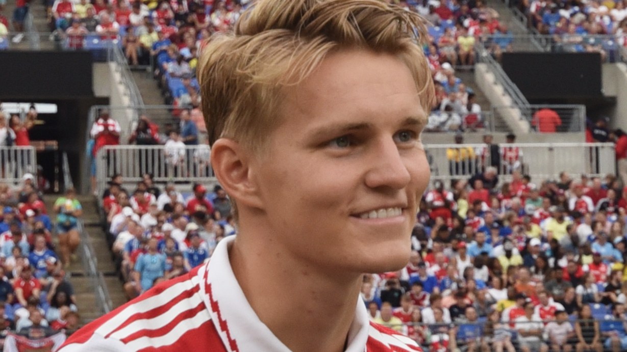 Young man with blond hair in a red-and-white sports jersey smiles during a crowded stadium event.