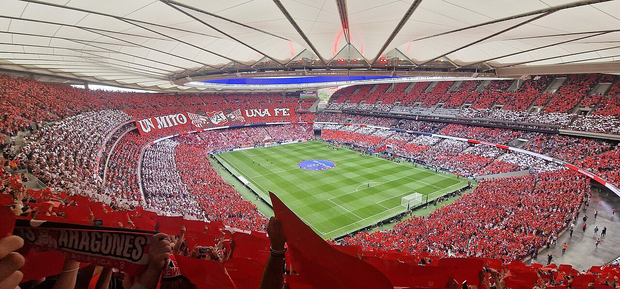 A packed soccer stadium filled with red and white supporters, banners waving above the field during a match.
