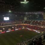 Nighttime stadium filled with fans holding red lights, large screen at left and illuminated field below.