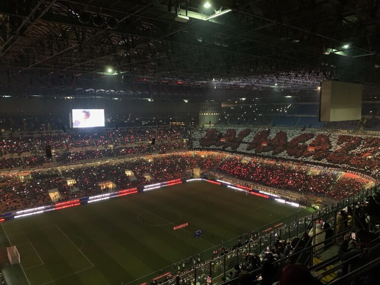 Nighttime stadium filled with fans holding red lights, large screen at left and illuminated field below.