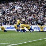 Soccer team in yellow jerseys celebrates on the field as fans cheer in the stands behind them.