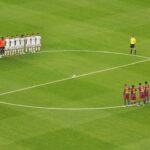 Two football teams stand in line facing each other around the center circle on a green pitch, with a referee nearby and the ball at the center.