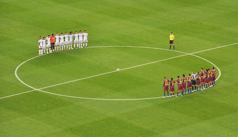 Two football teams stand in line facing each other around the center circle on a green pitch, with a referee nearby and the ball at the center.