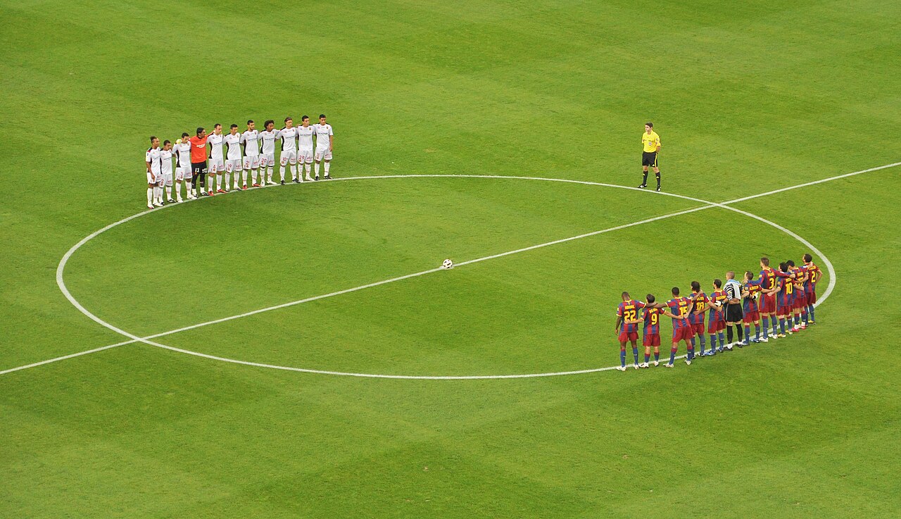 Two football teams stand in line facing each other around the center circle on a green pitch, with a referee nearby and the ball at the center.