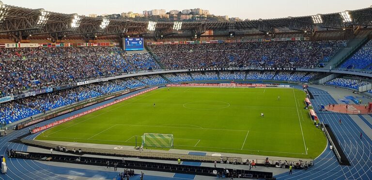 Stadium filled with blue seats, a green soccer pitch, and a running track around the field with a goal in the foreground.