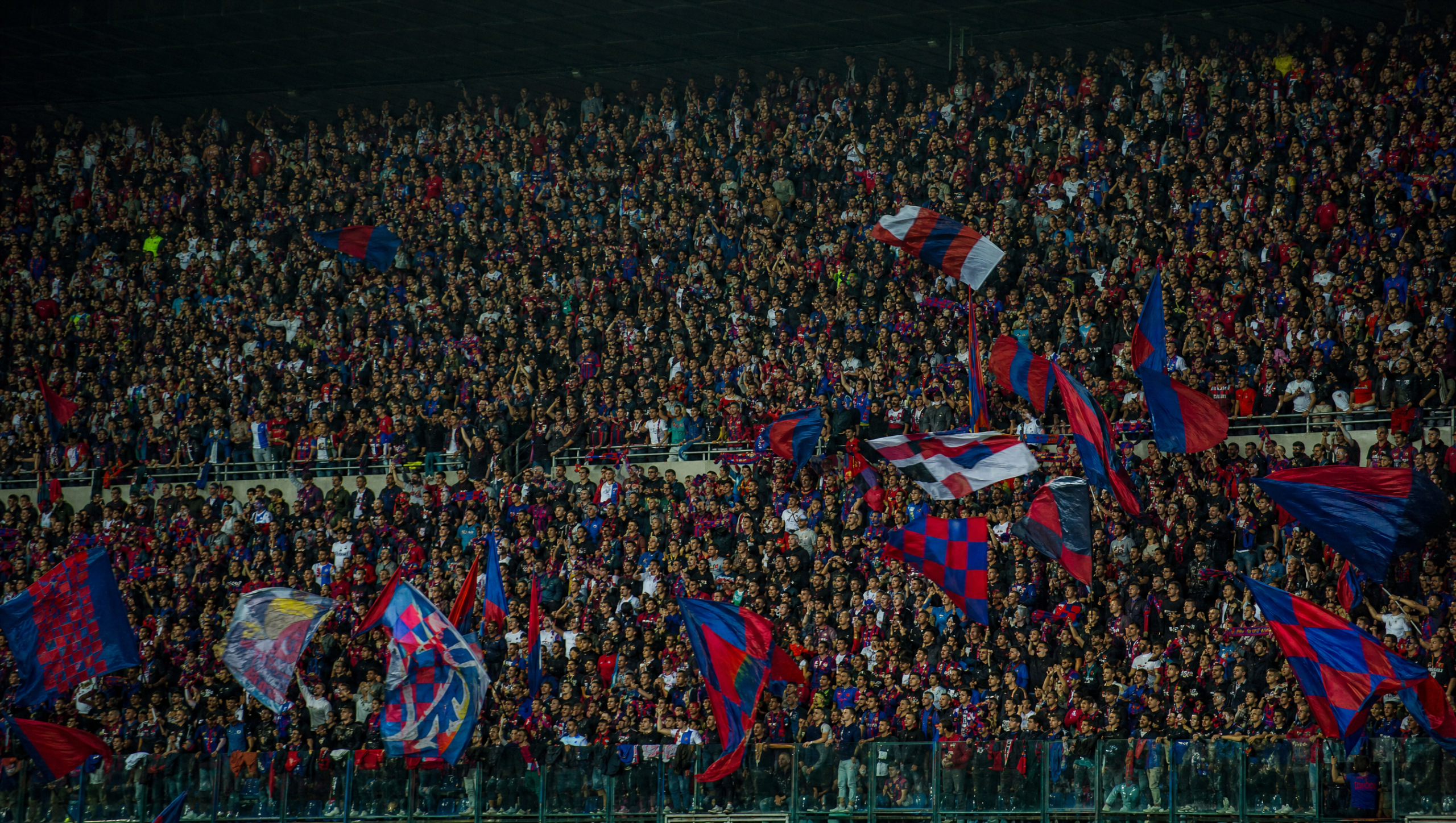 Dense crowd in a stadium stands waving red, blue, and white flags at a sporting event