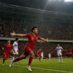 Soccer player in red celebrates a goal with arms wide as the stadium crowd cheers in the background.