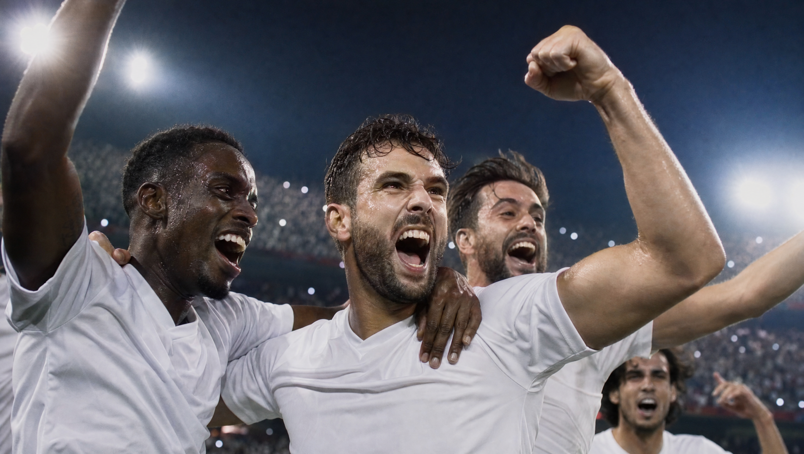 Soccer teammates celebrate a victory, arms around each other, on the field under stadium lights at night.